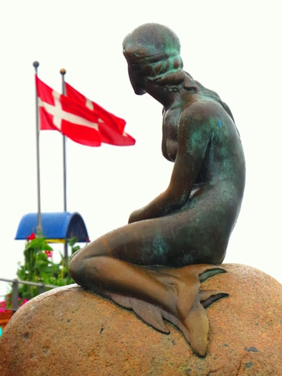 A bronze statue of a mermaid sitting on a rock, with two red and white flags in the background. The mermaid has her head turned, with detailed braids and fins, conveying a sense of contemplation. The background features some greenery and a blue structure.