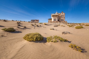 A large, elegant house stands amidst a desert landscape with sand dunes and sparse vegetation. The sky is clear and blue, enhancing the serenity of the scene. Another building, partially in ruins, is visible in the background, adding a sense of abandonment.