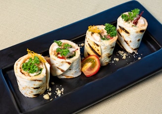 Four pieces of grilled wraps filled with cream and garnished with green herbs are neatly arranged on a rectangular black plate. A halved cherry tomato sits next to the wraps, adding a pop of color to the dish. Crumbs are scattered around the wraps for a textured presentation.