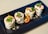 Four pieces of grilled wraps filled with cream and garnished with green herbs are neatly arranged on a rectangular black plate. A halved cherry tomato sits next to the wraps, adding a pop of color to the dish. Crumbs are scattered around the wraps for a textured presentation.
