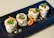 Four pieces of grilled wraps filled with cream and garnished with green herbs are neatly arranged on a rectangular black plate. A halved cherry tomato sits next to the wraps, adding a pop of color to the dish. Crumbs are scattered around the wraps for a textured presentation.