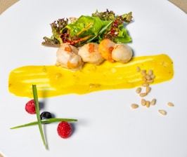 A gourmet dish is artistically presented on a white plate. Scallops, garnished with a bright orange sauce, are accompanied by a leafy green salad. A smear of vibrant yellow sauce runs across the plate, and scattered pine nuts add texture. Two red raspberries and a blueberry are positioned artfully alongside two thin green chives.