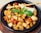 Chunks of grilled chicken are mixed with vibrant red and yellow bell peppers, garnished with fresh basil leaves and a sprinkle of herbs. A thin breadstick and a wedge of cheese are placed on top of the dish, served in a black bowl.
