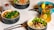 An assortment of colorful and healthy meals is arranged on a table. There are bowls containing salads with fresh vegetables like broccoli, peas, greens, and other garnishes. A glass of chia pudding with berries is placed alongside a refreshing beverage garnished with citrus fruits and mint leaves.