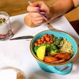 A colorful bowl containing quinoa, broccoli, chickpeas, sautéed spinach, sweet potato slices, and a lemon wedge. A hand with purple nail polish holds a fork poised to take a bite. On the table, there is a knife, a glass with chia pudding topped with strawberries and seeds, and a piece of bread.