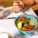 A colorful bowl containing quinoa, broccoli, chickpeas, sautéed spinach, sweet potato slices, and a lemon wedge. A hand with purple nail polish holds a fork poised to take a bite. On the table, there is a knife, a glass with chia pudding topped with strawberries and seeds, and a piece of bread.