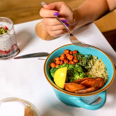 A colorful bowl containing quinoa, broccoli, chickpeas, sautéed spinach, sweet potato slices, and a lemon wedge. A hand with purple nail polish holds a fork poised to take a bite. On the table, there is a knife, a glass with chia pudding topped with strawberries and seeds, and a piece of bread.