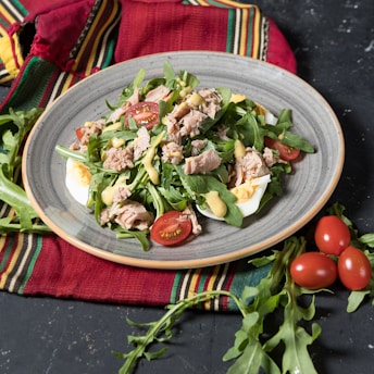 A plate of fresh salad containing arugula, cherry tomatoes, tuna chunks, and sliced boiled eggs, dressed with a mustard sauce. The salad is placed on a grayish plate, which rests on a multicolored cloth with red, green, yellow, and black stripes. Several whole cherry tomatoes and arugula leaves are scattered around the plate on a dark surface.