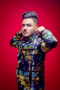 A person with styled hair is wearing a colorful jacket with a graffiti-like pattern and the word 'Nike' printed multiple times. The individual stands against a vivid red background, adjusting the jacket's collar.