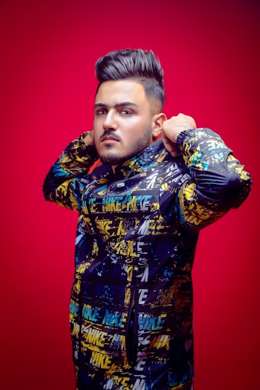 A person with styled hair is wearing a colorful jacket with a graffiti-like pattern and the word 'Nike' printed multiple times. The individual stands against a vivid red background, adjusting the jacket's collar.