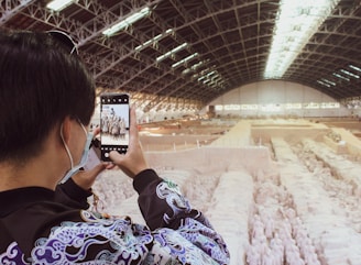 A person wearing a patterned jacket and a face mask is taking a photo with a smartphone. The background features an expansive indoor archaeological site with rows of ancient terracotta warrior statues. The site is housed under a large, arched ceiling with structural beams and skylights.