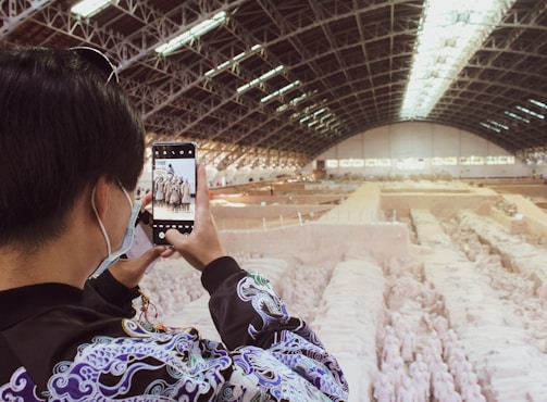 A person wearing a patterned jacket and a face mask is taking a photo with a smartphone. The background features an expansive indoor archaeological site with rows of ancient terracotta warrior statues. The site is housed under a large, arched ceiling with structural beams and skylights.