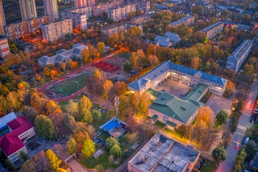 An aerial view of a city area featuring residential buildings, a central school building with a u-shaped layout, and sports fields. The scene is filled with colorful autumn trees, providing a vibrant contrast to the urban structures. The streets are illuminated with streetlights, indicating an evening setting.