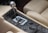 A close-up view of a car's manual gear shift lever positioned within a polished metallic gate. Surrounding the gear lever are multiple control buttons and dials, possibly for climate and other in-car functionalities. The interior is upholstered with beige leather featuring detailed stitching.