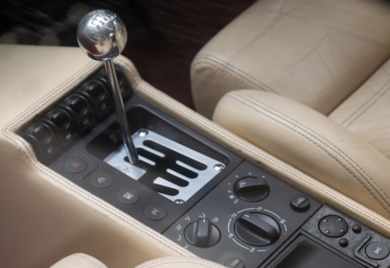 A close-up view of a car's manual gear shift lever positioned within a polished metallic gate. Surrounding the gear lever are multiple control buttons and dials, possibly for climate and other in-car functionalities. The interior is upholstered with beige leather featuring detailed stitching.