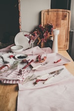 A rustic kitchen scene featuring dried red chili peppers arranged on a wooden surface. There is a cutting board standing vertically in the background with a clear jar next to it. A white salt or pepper grinder is placed on the right side. A cocktail glass holds some of the dried peppers. Various kitchen utensils, including a fork and knife with white handles, are laid out alongside a white plate, which is partially obscured by a striped cloth. A small white bowl filled with dark, circular spices or small fruits is visible.