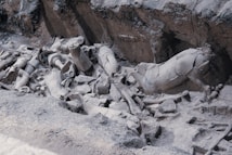 Fragments of ancient statues and sculptures are scattered across a rocky excavation site. The pieces appear timeworn and dusty, with some recognizable parts like limbs and torsos visible among the debris.