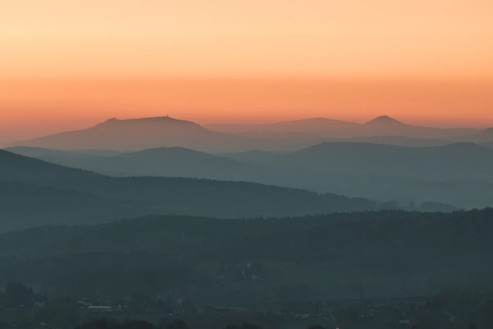 The image depicts a breathtaking view of a mountain range at sunset. The mountains, painted in shades of orange and pink, stretch across the horizon, creating a stunning backdrop for the sky. The sun is setting behind these majestic peaks, casting long shadows that add depth to the scene. In the foreground, there are trees scattered throughout the landscape, their green foliage contrasting with the warm hues of the sunset. The image is taken from a high vantage point, providing an expansive view of the mountains and the surrounding area.