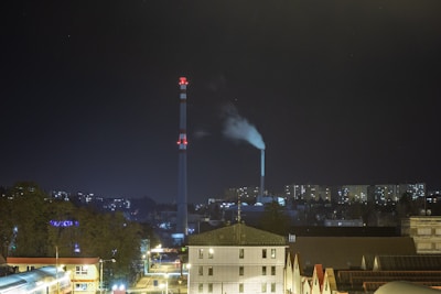 A cityscape at night featuring illuminated buildings and two tall chimneys with red warning lights. The larger chimney emits white smoke against a dark sky, and residential buildings with lit windows are visible in the background.
