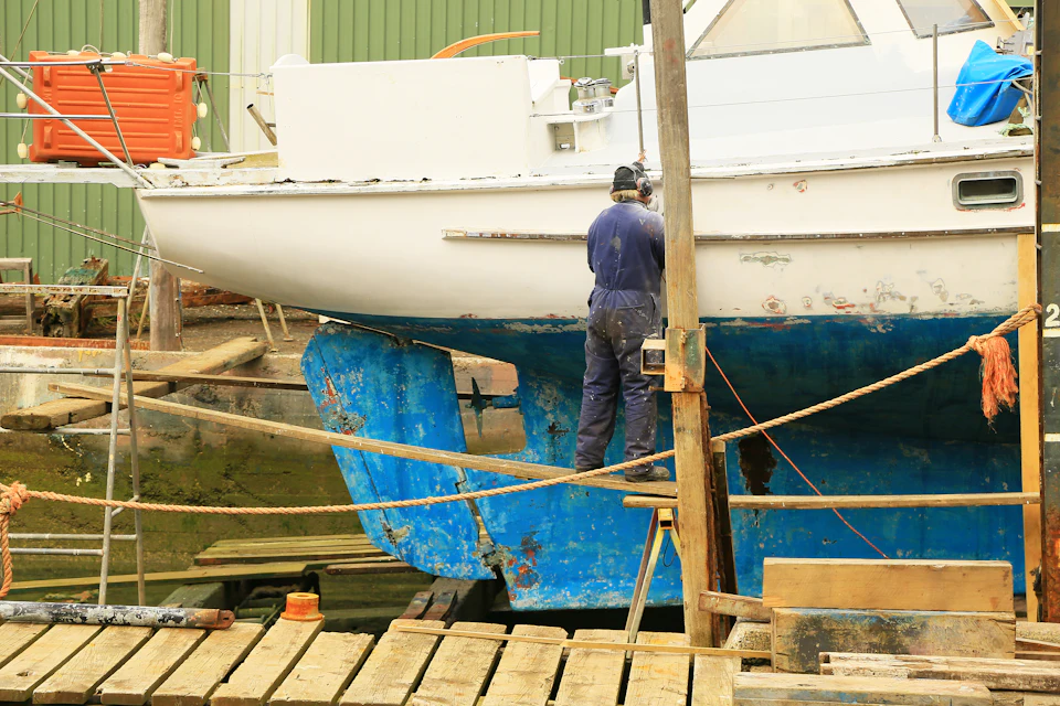 Worker repairing the hull of a white and blue boat on a slipway
