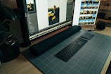 A desktop workspace featuring a large monitor displaying photo editing software. The arrangement includes a stylish black keyboard and a sleek digital drawing pad resting on a grid-patterned mat. To the side, a camera sits on the wooden desk, and there are plants adding a touch of greenery.