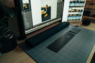 A desktop workspace featuring a large monitor displaying photo editing software. The arrangement includes a stylish black keyboard and a sleek digital drawing pad resting on a grid-patterned mat. To the side, a camera sits on the wooden desk, and there are plants adding a touch of greenery.