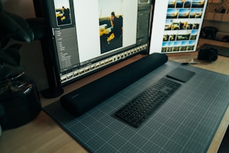 A desktop workspace featuring a large monitor displaying photo editing software. The arrangement includes a stylish black keyboard and a sleek digital drawing pad resting on a grid-patterned mat. To the side, a camera sits on the wooden desk, and there are plants adding a touch of greenery.