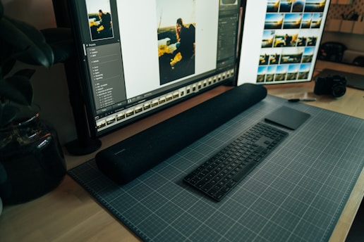 A desktop workspace featuring a large monitor displaying photo editing software. The arrangement includes a stylish black keyboard and a sleek digital drawing pad resting on a grid-patterned mat. To the side, a camera sits on the wooden desk, and there are plants adding a touch of greenery.