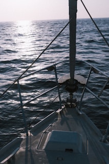 A sailboat deck facing towards the open sea with gentle waves and a reflection of sunlight on the water surface. The boat is equipped with a sturdy railing along the sides and a mast in the front.