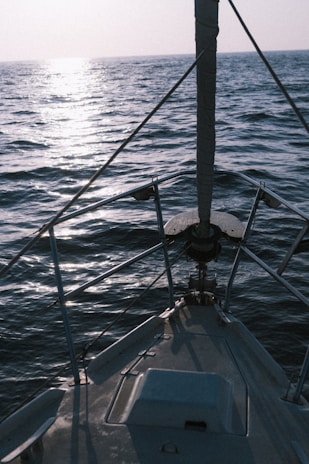 A sailboat deck facing towards the open sea with gentle waves and a reflection of sunlight on the water surface. The boat is equipped with a sturdy railing along the sides and a mast in the front.