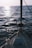 A sailboat deck facing towards the open sea with gentle waves and a reflection of sunlight on the water surface. The boat is equipped with a sturdy railing along the sides and a mast in the front.