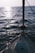 A sailboat deck facing towards the open sea with gentle waves and a reflection of sunlight on the water surface. The boat is equipped with a sturdy railing along the sides and a mast in the front.