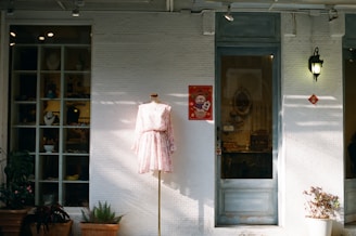 A storefront with a mannequin displaying a light pink floral dress is positioned in front of a white brick wall. To the side, there is a large window showcasing various items, possibly jewelry or small decorations. A poster with a cartoon character hangs next to a door with a vintage handle. Plants in pots are placed at the base of the wall, providing a touch of greenery. A lantern-style light fixture is mounted on the wall, casting a soft glow.