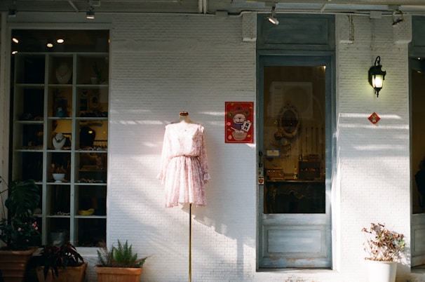 A storefront with a mannequin displaying a light pink floral dress is positioned in front of a white brick wall. To the side, there is a large window showcasing various items, possibly jewelry or small decorations. A poster with a cartoon character hangs next to a door with a vintage handle. Plants in pots are placed at the base of the wall, providing a touch of greenery. A lantern-style light fixture is mounted on the wall, casting a soft glow.
