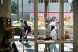 A view from inside a shop looking out through a large window. Jewelry and small decorative items are displayed on a table by the window. Outside, several people are walking along the street, with some wearing masks. The background features greenery and a colorful mural on the wall.