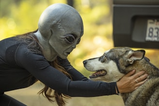 A person wearing an alien mask is gently holding the head of a dog, looking closely into its eyes. The background is blurred with hints of greenery, and part of a vehicle with a visible license plate is on the right side.