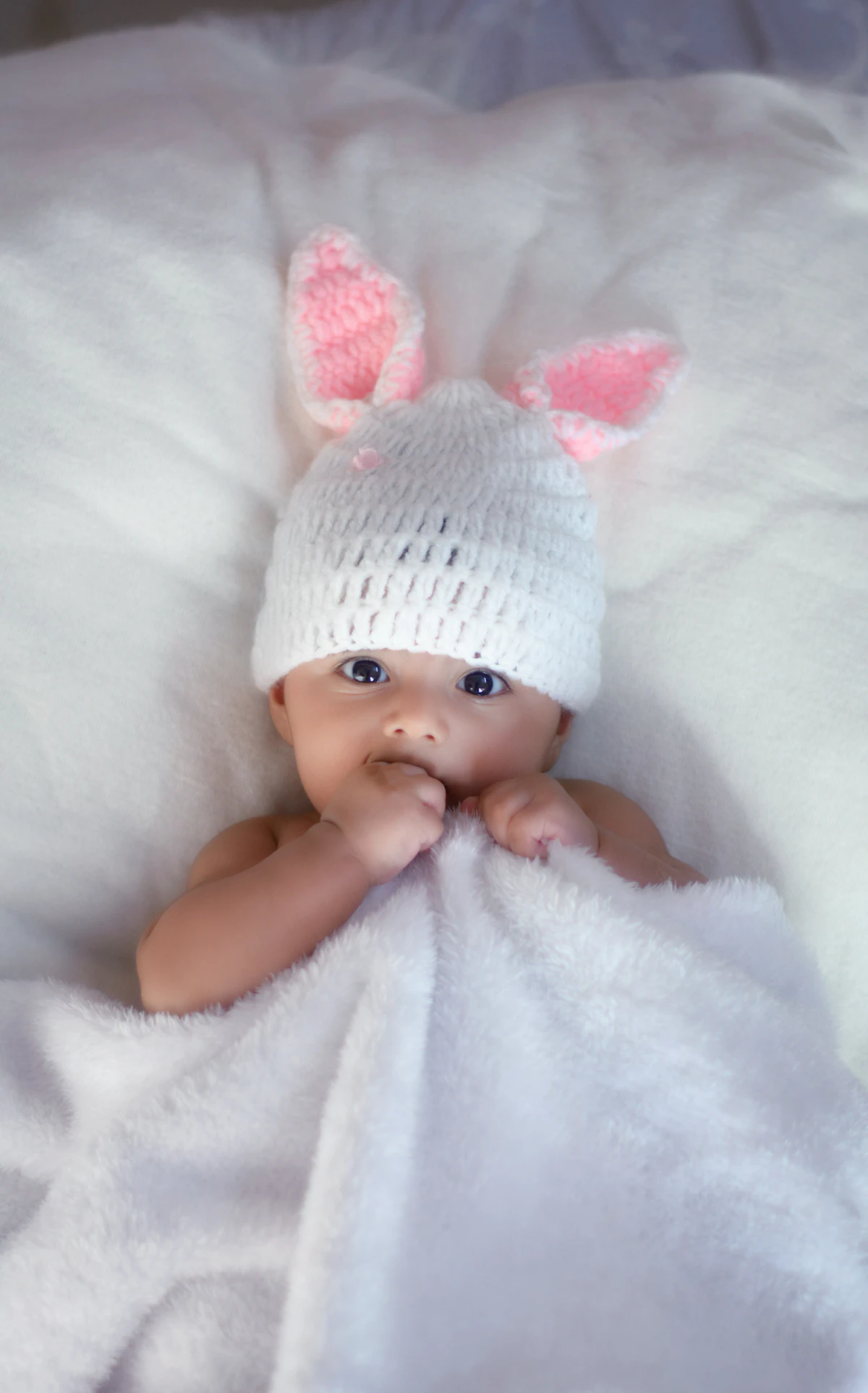 Newborn baby wearing a white knit cap lying on their back on soft white bedding