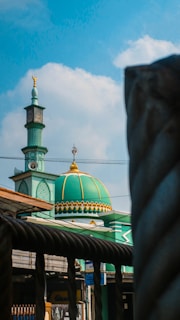 A mosque with a green dome and minaret against a blue sky with scattered clouds. The dome features golden accents and an intricate design. Foreground includes a metal fence and some cables.