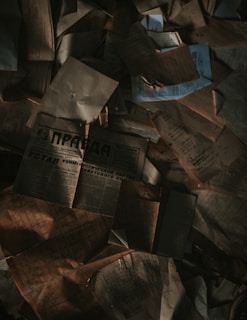 A collection of old, weathered documents and newspapers scattered in a disorganized manner. The papers are crumpled, and some have shadowy areas creating a dim, mysterious atmosphere.