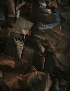 A collection of old, weathered documents and newspapers scattered in a disorganized manner. The papers are crumpled, and some have shadowy areas creating a dim, mysterious atmosphere.