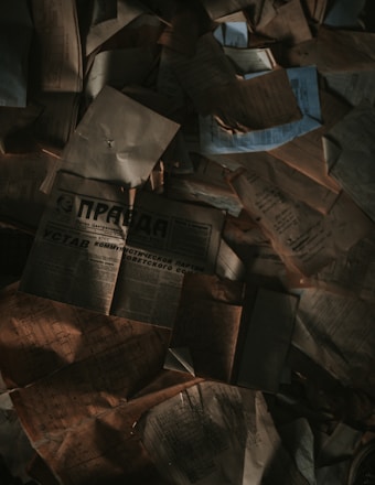 A collection of old, weathered documents and newspapers scattered in a disorganized manner. The papers are crumpled, and some have shadowy areas creating a dim, mysterious atmosphere.