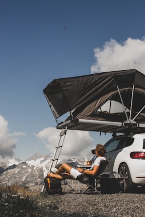 A person is sitting comfortably in a folding chair beside a white car, which has a rooftop tent set up with a ladder. The scene takes place on a gravel surface overlooking a mountainous landscape on a clear day with some clouds in the sky.