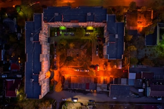 Aerial view of a large, L-shaped apartment building enclosing a central courtyard. The area is illuminated by vibrant street lights casting orange and green hues. Several cars are parked along the building perimeter and in the courtyard. Adjacent structures and surrounding trees are visible, with the scene captured during twilight or nighttime.