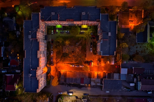 Aerial view of a large, L-shaped apartment building enclosing a central courtyard. The area is illuminated by vibrant street lights casting orange and green hues. Several cars are parked along the building perimeter and in the courtyard. Adjacent structures and surrounding trees are visible, with the scene captured during twilight or nighttime.