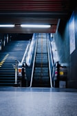 An empty escalator rises between two static railings with sleek metal sides, leading upwards in a dimly lit underground station. Adjacent to the left, a set of stairs is visible, illuminated by overhead fluorescent lights. The space is marked by industrial design elements with a cold, metallic feel.