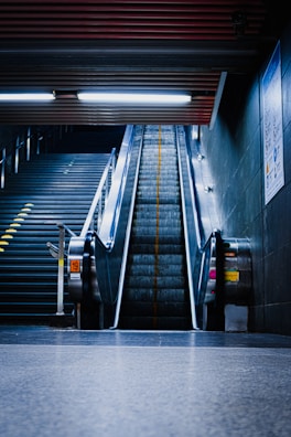 An empty escalator rises between two static railings with sleek metal sides, leading upwards in a dimly lit underground station. Adjacent to the left, a set of stairs is visible, illuminated by overhead fluorescent lights. The space is marked by industrial design elements with a cold, metallic feel.
