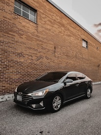 A sleek black sedan is parked next to a large brick wall. The car's headlights are on, and the windows are tinted. The wall has two windows and is made of reddish-brown bricks, and the ground is surfaced with asphalt.
