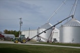 A green tractor is parked on a gravel path near a farm building. Adjacent to the tractor, there is an agricultural auger conveyor connected to large silver grain silos. Several power lines and a utility pole are visible, with grassy areas surrounding the scene.