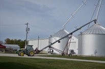 A green tractor is parked on a gravel path near a farm building. Adjacent to the tractor, there is an agricultural auger conveyor connected to large silver grain silos. Several power lines and a utility pole are visible, with grassy areas surrounding the scene.