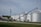 A green tractor is parked on a gravel path near a farm building. Adjacent to the tractor, there is an agricultural auger conveyor connected to large silver grain silos. Several power lines and a utility pole are visible, with grassy areas surrounding the scene.